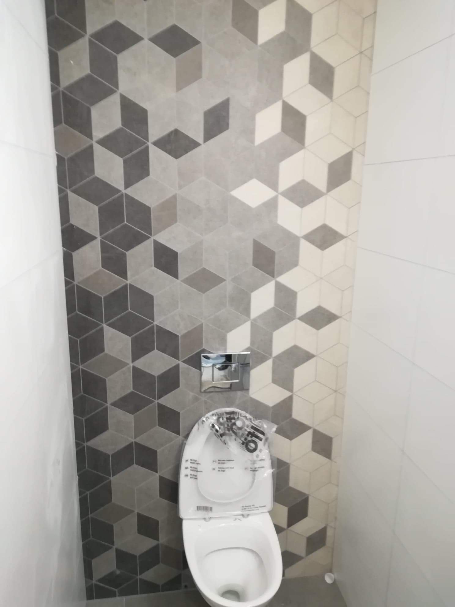 Zamontowana toaleta podwieszana z chromowanym przyciskiem spłukującym na ścianie wyłożonej płytkami w geometryczny wzór 3D w odcieniach szarości i bieli, z folią ochronną na desce sedesowej.