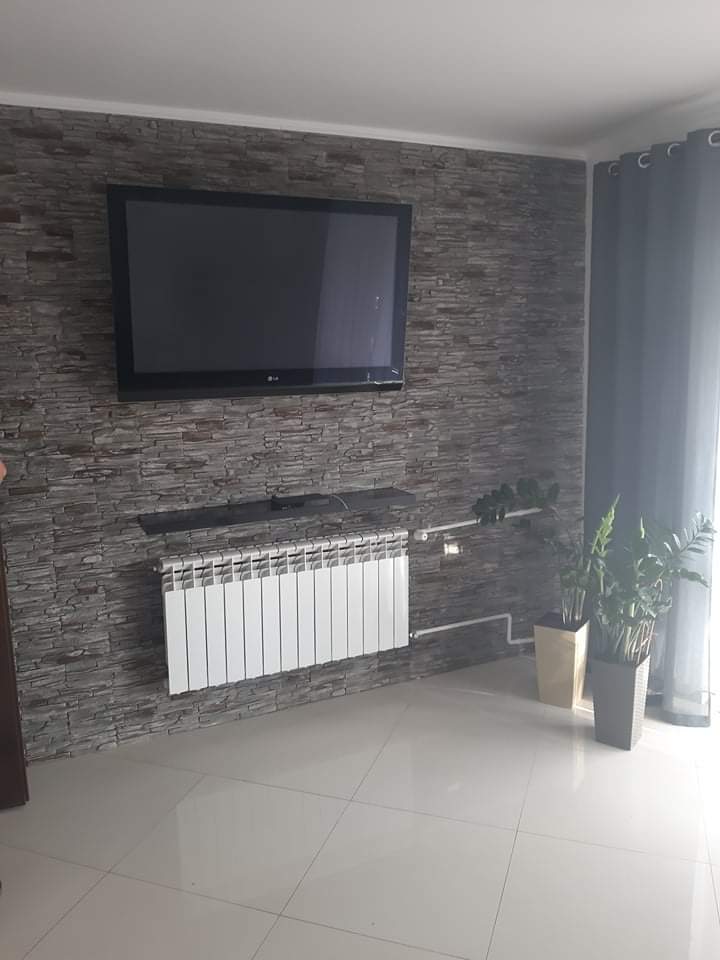 Telewizor na ścianie wyłożonej szarym kamieniem dekoracyjnym, biały grzejnik panelowy pod spodem, dwie doniczki z roślinami obok, jasna podłoga z płytek.