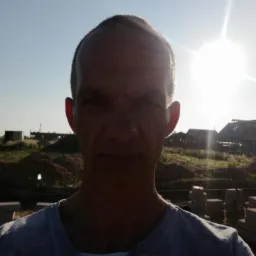 Mężczyzna na tle budowy w słoneczny dzień. Widoczne ułożone bloczki fundamentowe i częściowo zarys budynku. Silne światło słoneczne.