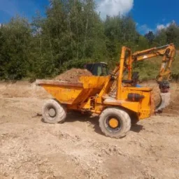 Żółta wywrotka z koparką na placu budowy, częściowo załadowana ziemią, widoczna na tle wykopu i zieleni.