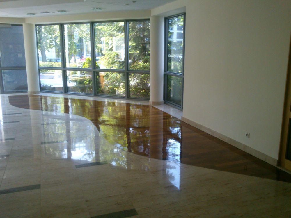 Błyszcząca podłoga w przestronnym wnętrzu z dużymi oknami, łącząca parkiet w odcieniach brązu i płytki w kolorze beżowym z ciemniejszymi akcentami, odbijająca światło dzienne.