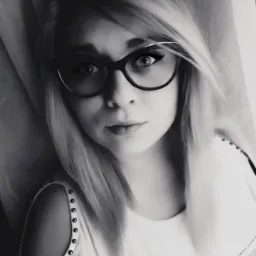 Portret młodej kobiety w okularach, z blond włosami i bluzką z ozdobnymi ćwiekami na ramieniu, w czarno-białej tonacji.