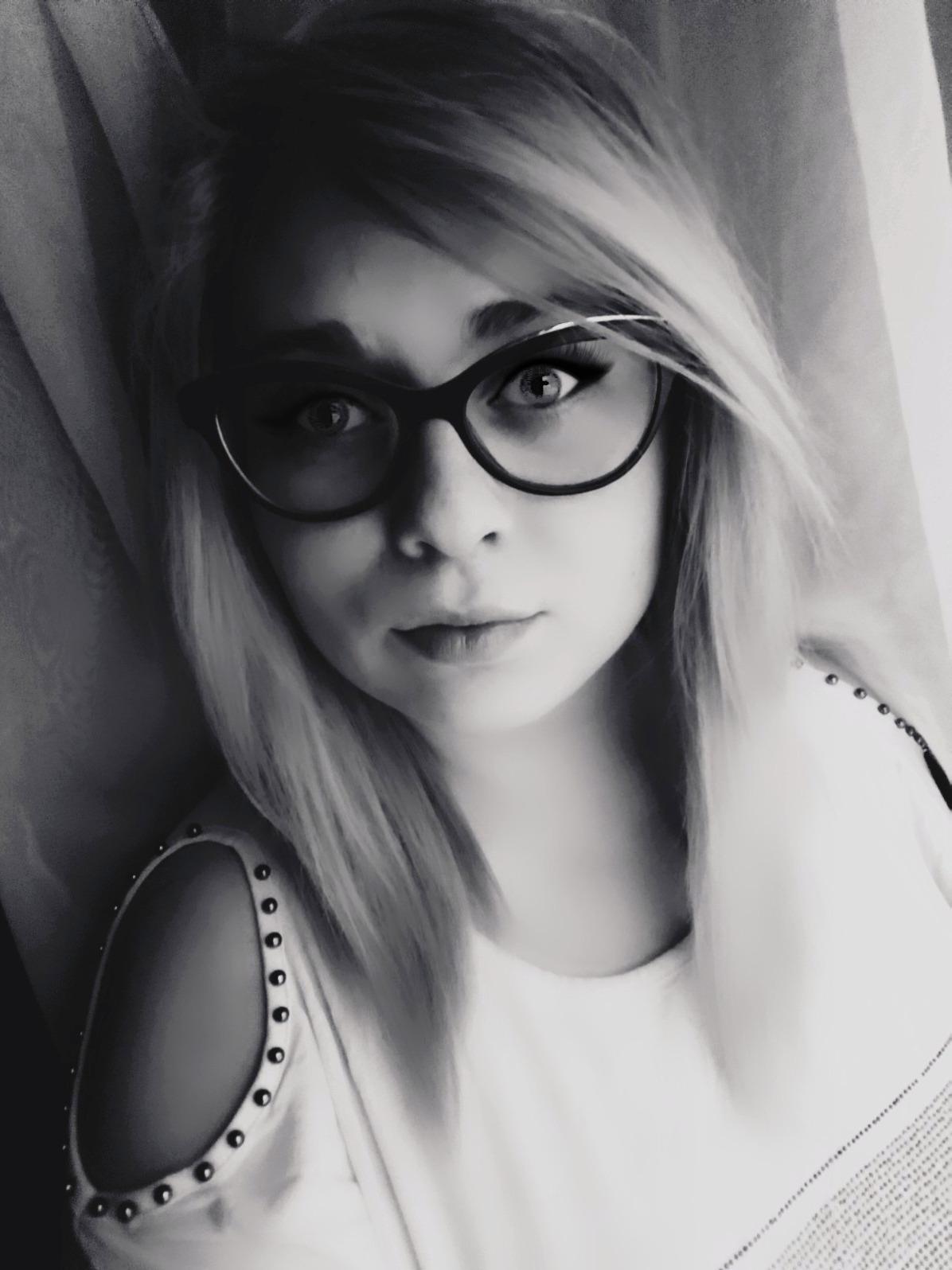 Portret młodej kobiety w okularach, z blond włosami i bluzką z ozdobnymi ćwiekami na ramieniu, w czarno-białej tonacji.