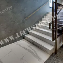 Nowoczesne schody wewnętrzne z białego marmuru z szarymi żyłami, z metalową balustradą z szybami, na tle szarej ściany z fakturą.