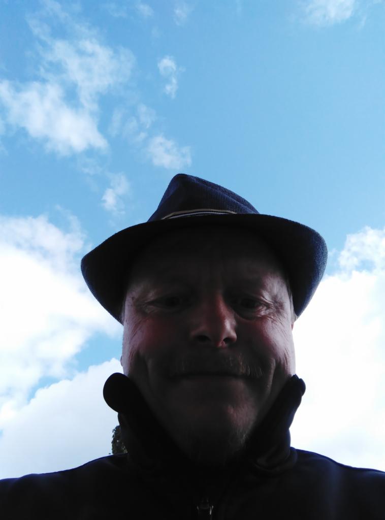 Portret mężczyzny w kapeluszu na tle błękitnego nieba z chmurami, ujęcie z dołu, oświetlenie naturalne.