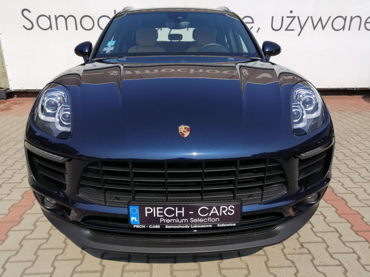 Granatowy samochód marki Porsche Macan, widok z przodu, z widocznym logo firmy Piech-Cars na tablicy rejestracyjnej i w tle napis 'Samochody używane'.