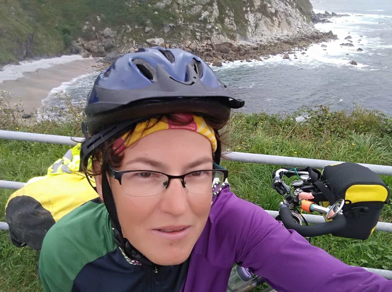 Uśmiechnięta kobieta w okularach i kasku rowerowym robiąca selfie na tle klifowego wybrzeża z widoczną plażą i morzem, w tle rower z sakwami.