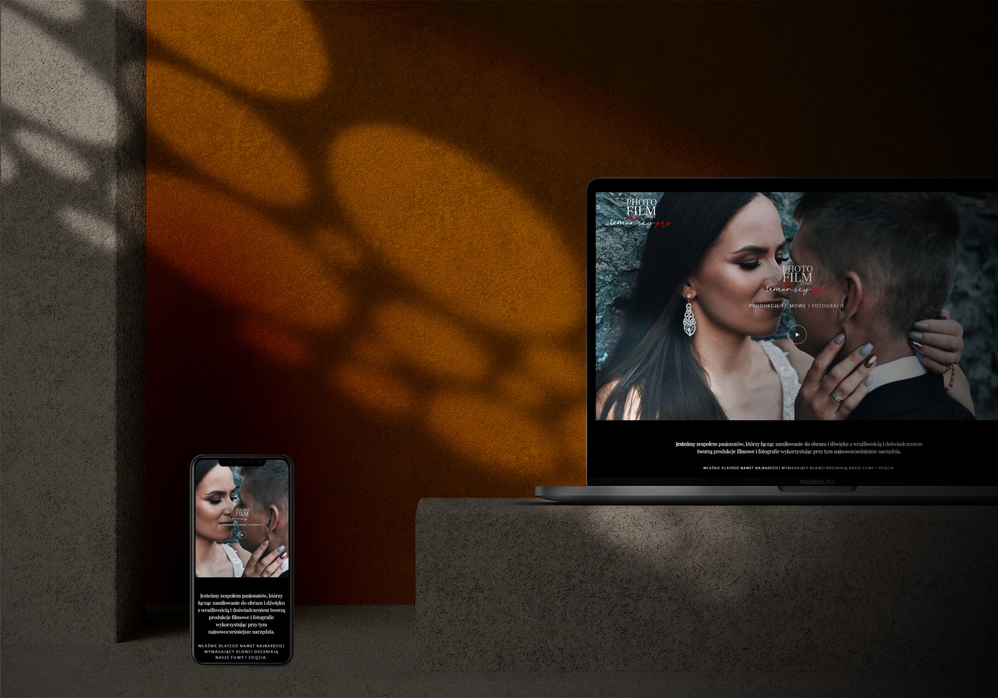 Laptop i smartfon prezentujące stronę internetową firmy fotograficzno-filmowej, z kadrem pary młodej na pierwszym planie, na tle abstrakcyjnej ściany w ciepłych barwach.