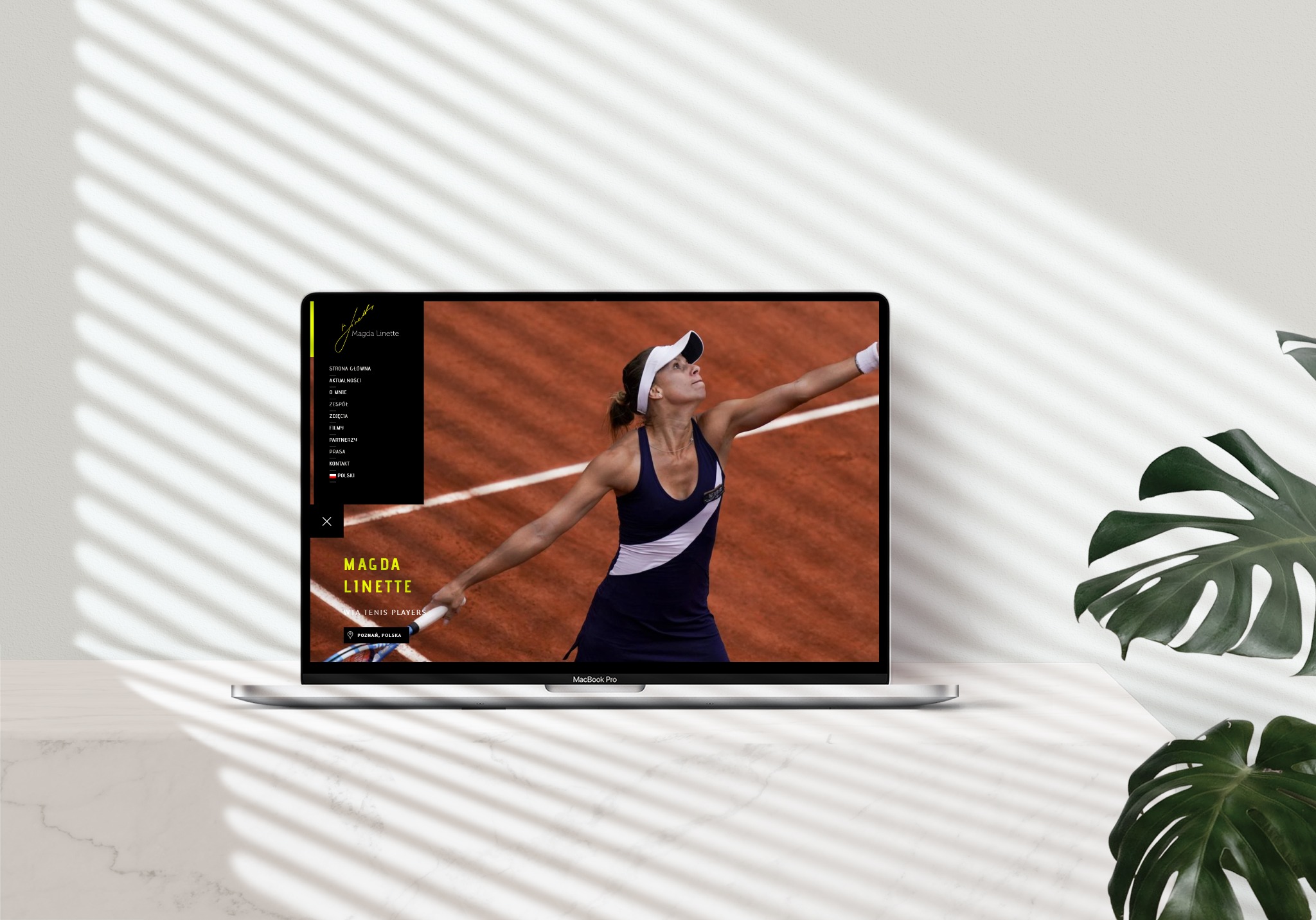 Laptop MacBook Pro wyświetlający stronę internetową tenisistki Magdy Linette na tle białego blatu z cieniami i liściem monstery.
