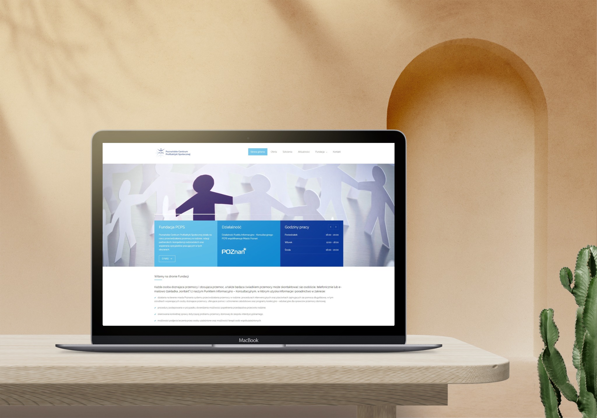Ekran laptopa MacBook wyświetlający stronę internetową Poznańskiego Centrum Profilaktyki Społecznej (PCPS) z grafiką papierowych postaci trzymających się za ręce, na beżowym biurku z kaktusem w tle.