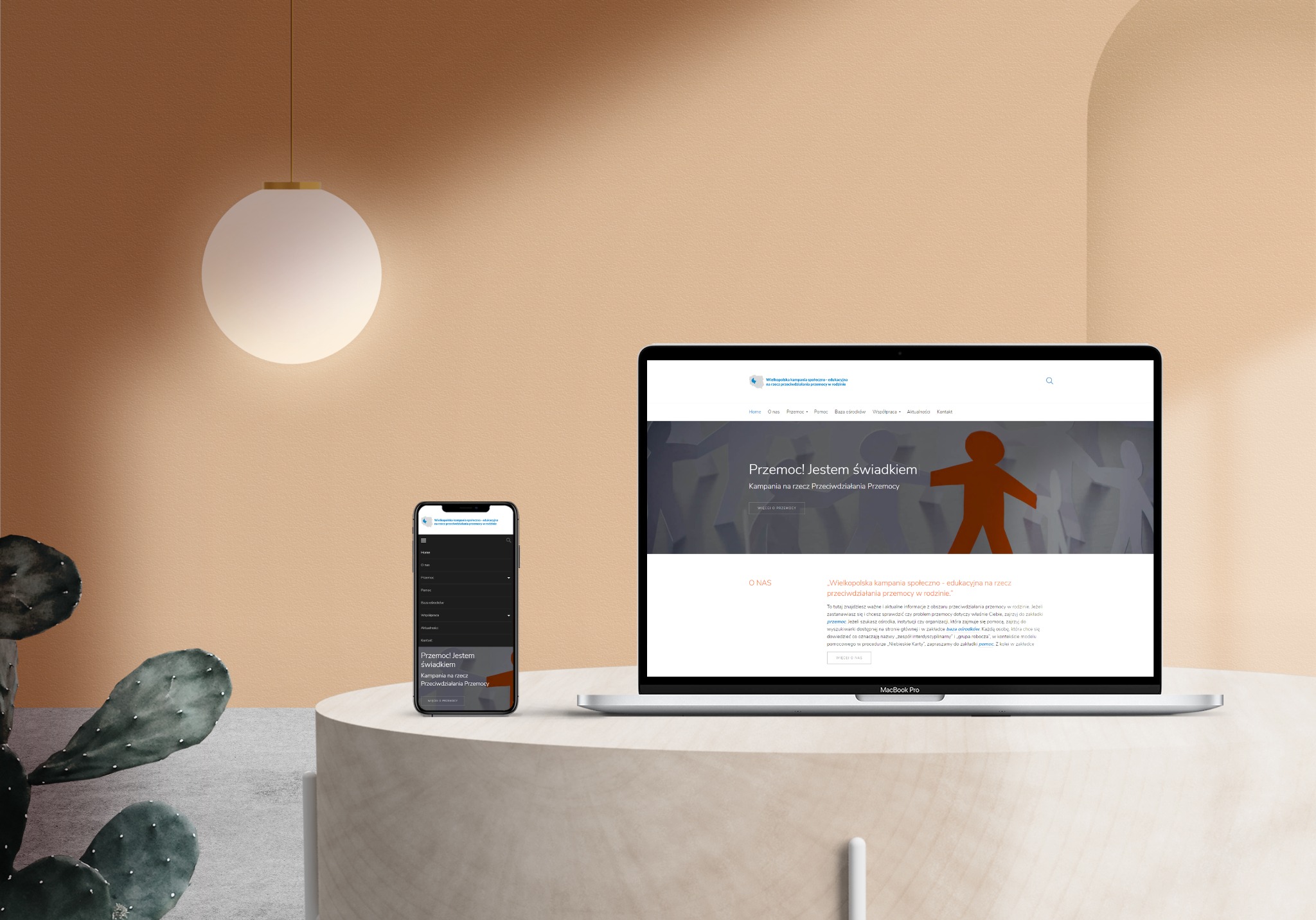 Ekran laptopa i smartfona wyświetlające stronę internetową kampanii społecznej 'Przemoc! Jestem świadkiem' na tle minimalistycznego wnętrza z kaktusem.