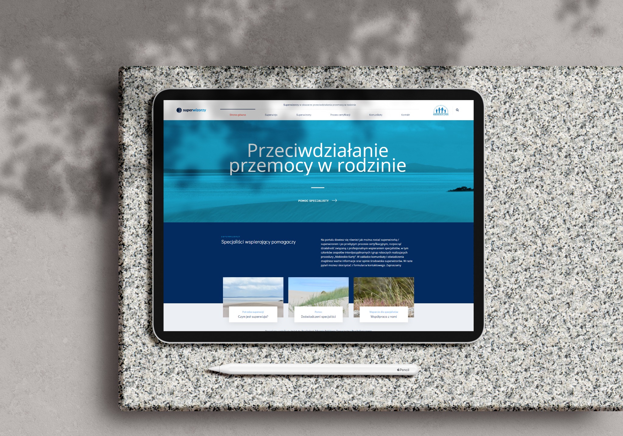 Tablet wyświetlający stronę internetową 'Superwizorzy' z hasłem 'Przeciwdziałanie przemocy w rodzinie', leżący na szarej, teksturowanej powierzchni obok białego rysika Apple Pencil.