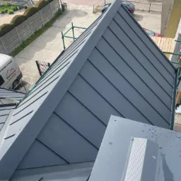 Szary, metalowy dach z widocznymi obróbkami blacharskimi i oknami dachowymi, sfotografowany z góry, z zielonymi rusztowaniami w tle.