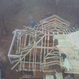 Eldach - Budowa domu w trakcie realizacji, widok z góry. Widoczne drewniane elementy konstrukcyjne dachu i ściany z cegieł. Na zewnątrz budynku leżą materiały budowlane.