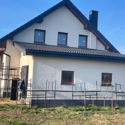 Eldach - Nowy dom jednorodzinny z ciemną dachówką i rynnami, widoczny fragment rusztowania. Jasna elewacja i okna w brązowych ramach, błękitne niebo w tle.