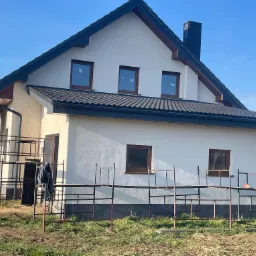 Nowy dom jednorodzinny z ciemną dachówką i rynnami, widoczny fragment rusztowania. Jasna elewacja i okna w brązowych ramach, błękitne niebo w tle.