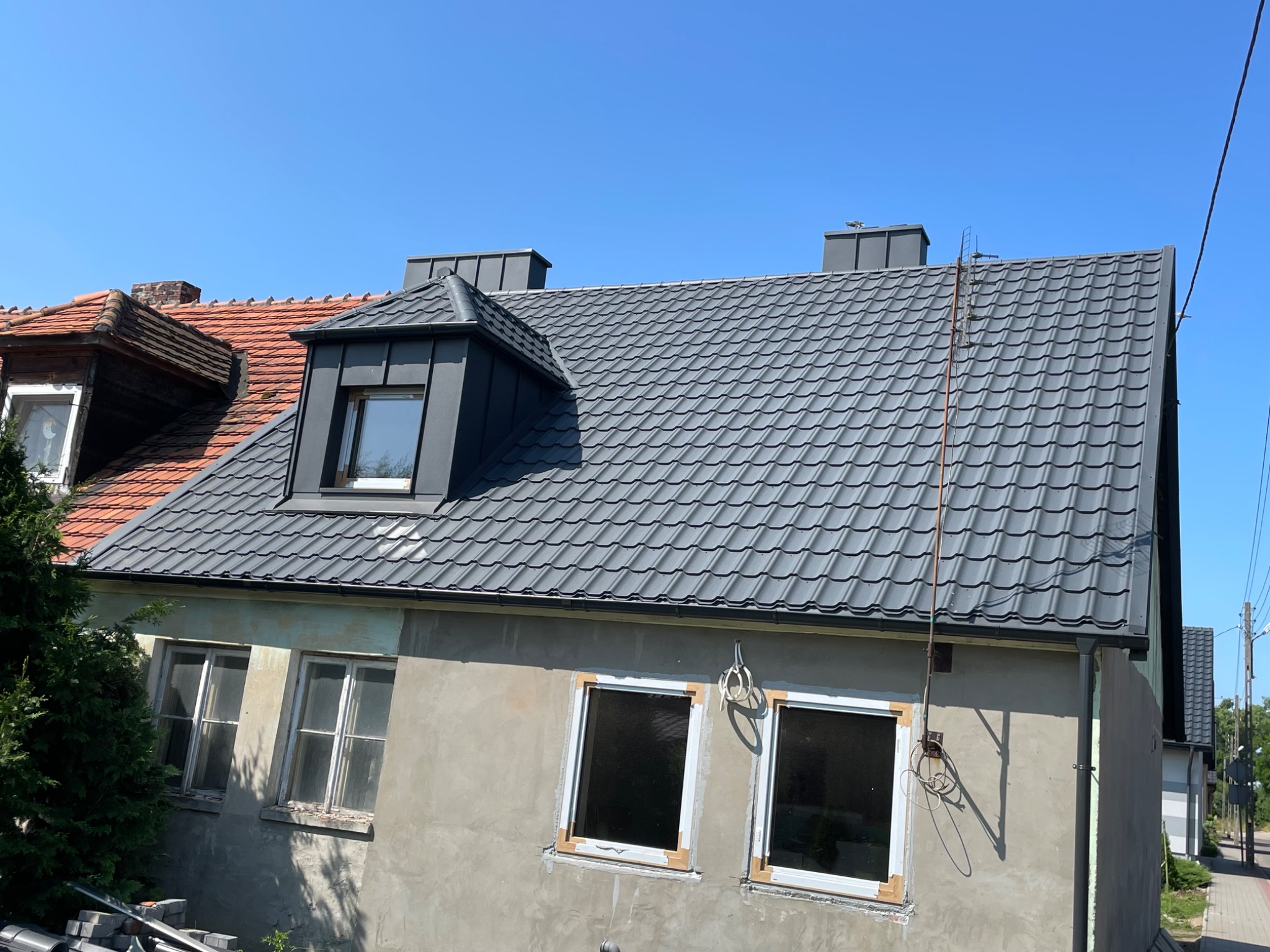 Szary dach z blachy trapezowej na budynku z oknami w trakcie montażu. Widoczne rynny i kominy. Część dachu w kolorze czerwonym. Błękitne niebo w tle.