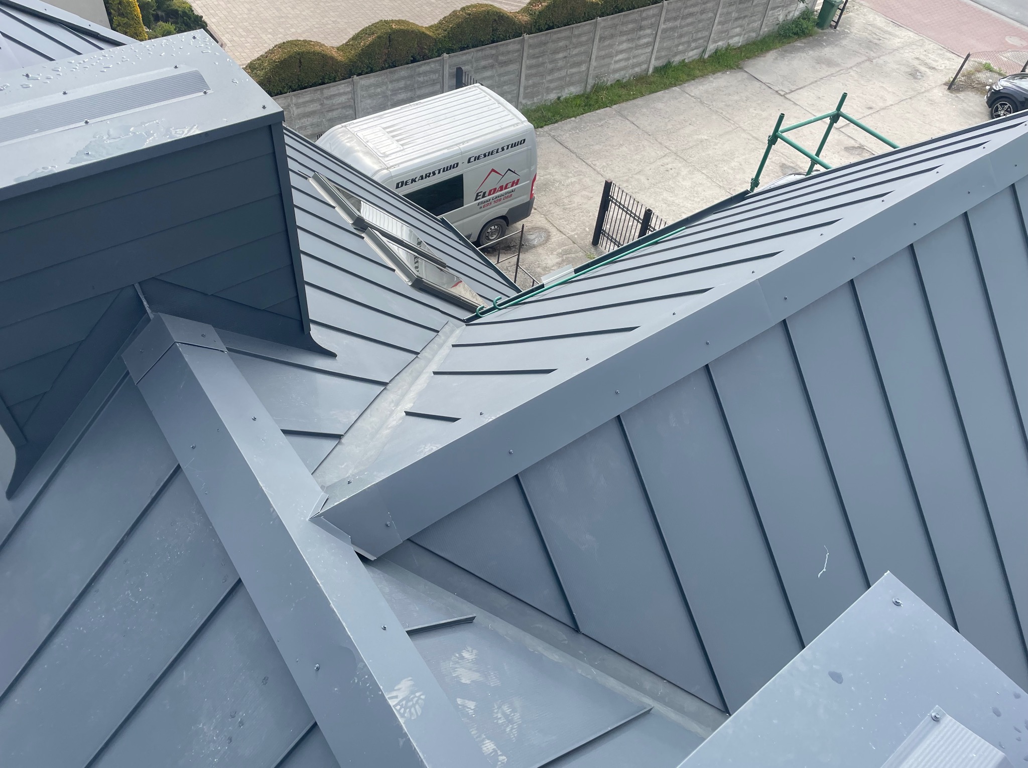 Szary, metalowy dach z widocznymi obróbkami blacharskimi i oknami dachowymi. W tle budynek i firmowy samochód dekarski.