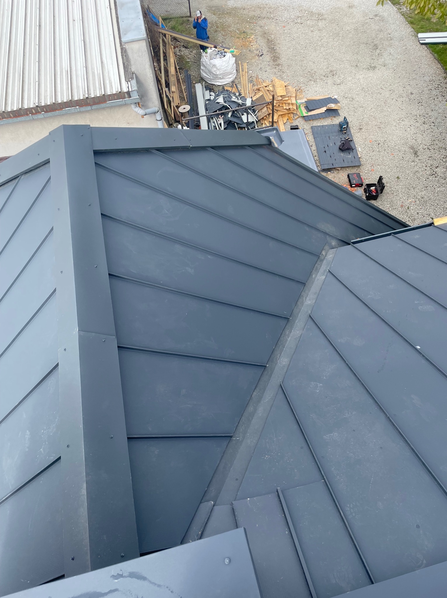 Szary dach z blachy trapezowej, widok z góry. Na dachu widoczne obróbki blacharskie i narzędzia dekarskie. W tle budynek i osoba w niebieskim ubraniu.