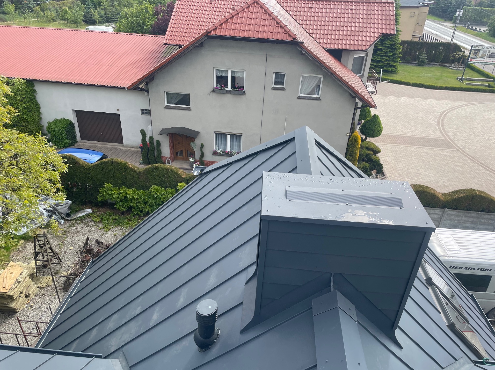 Nowoczesny, grafitowy dach z blachy na domu jednorodzinnym, widok z góry, elementy wentylacyjne, w tle budynki mieszkalne i zieleń. Blacha trapezowa.