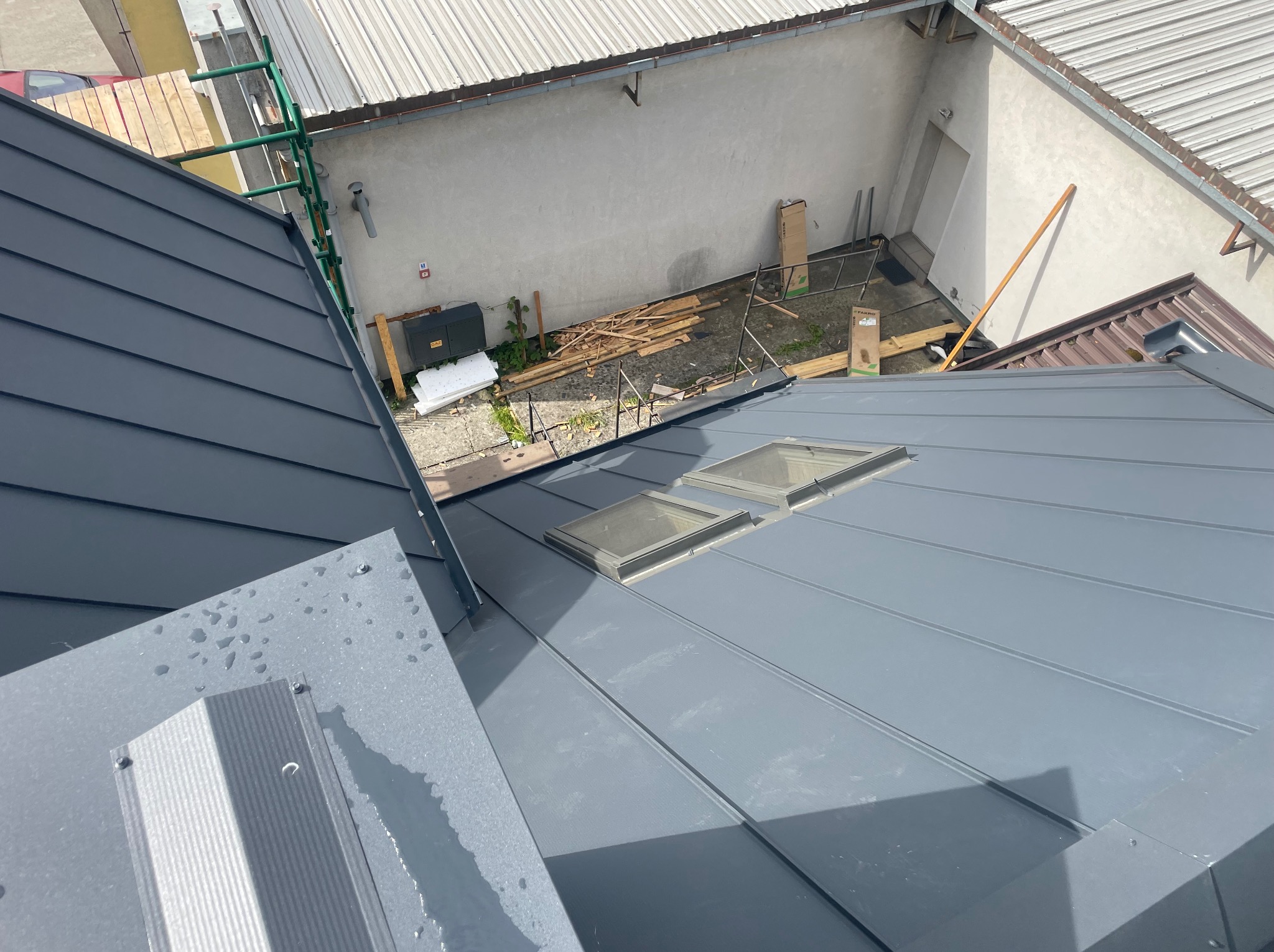 Szary, blaszany dach z dwoma oknami dachowymi i elementami obróbki blacharskiej, sfotografowany z góry w słoneczny dzień, z widocznymi rusztowaniami w tle.