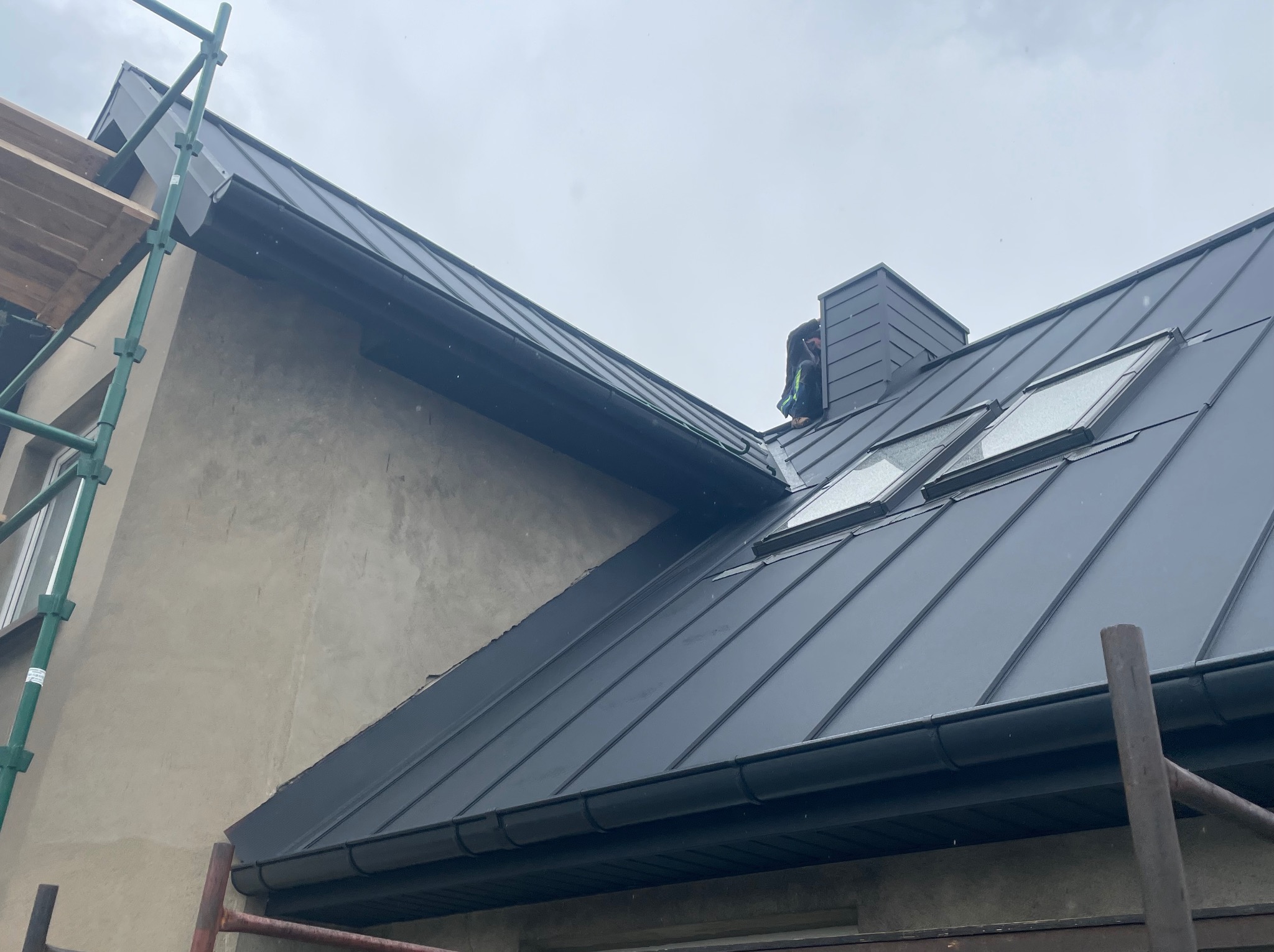 Czarny dach z blachy na domu jednorodzinnym, widoczne okna dachowe, komin i rynny. W tle szare niebo i fragment rusztowania. Prace dekarskie w toku.