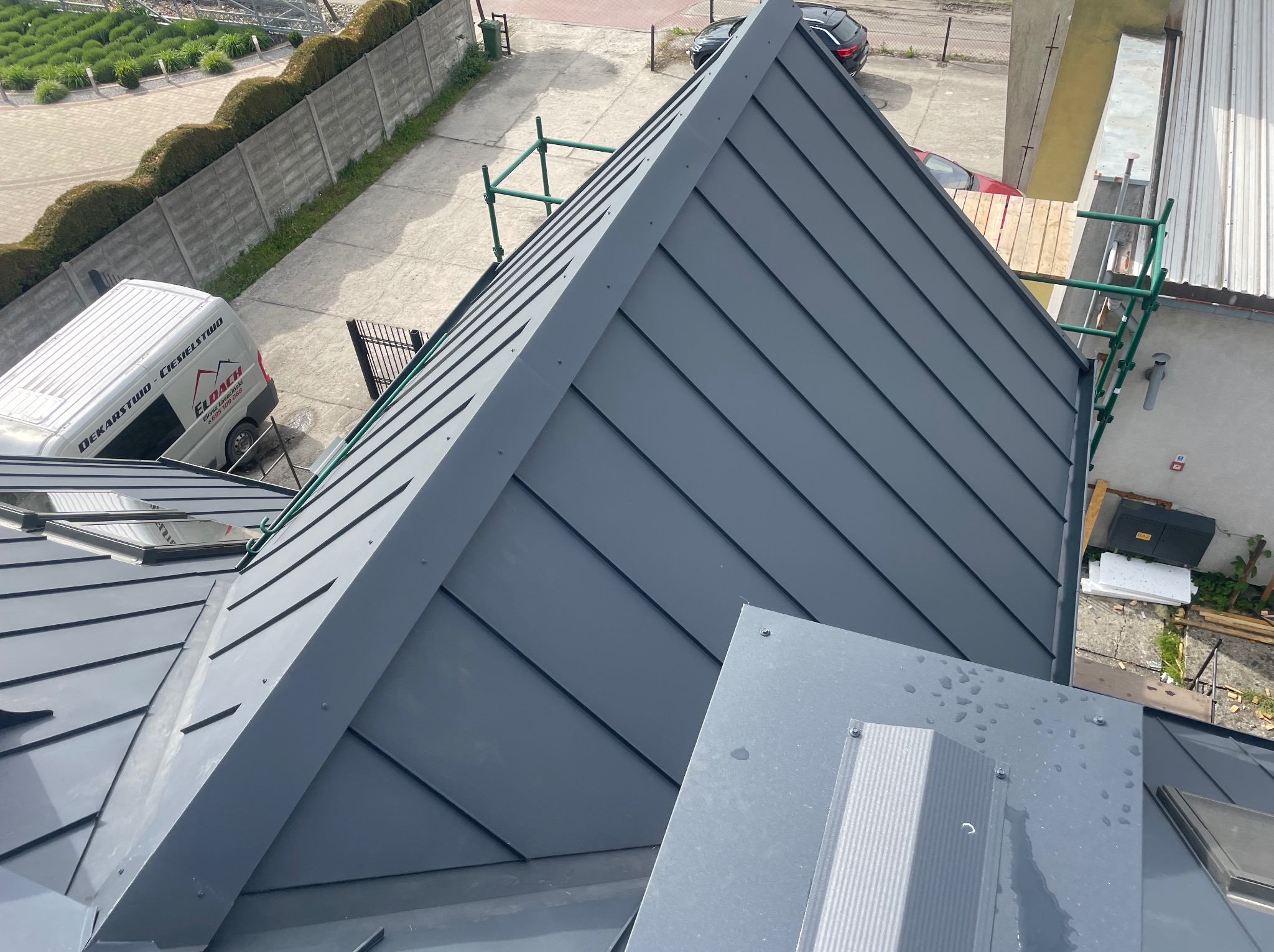 Szary, metalowy dach z widocznymi obróbkami blacharskimi i oknami dachowymi, sfotografowany z góry, z zielonymi rusztowaniami w tle.