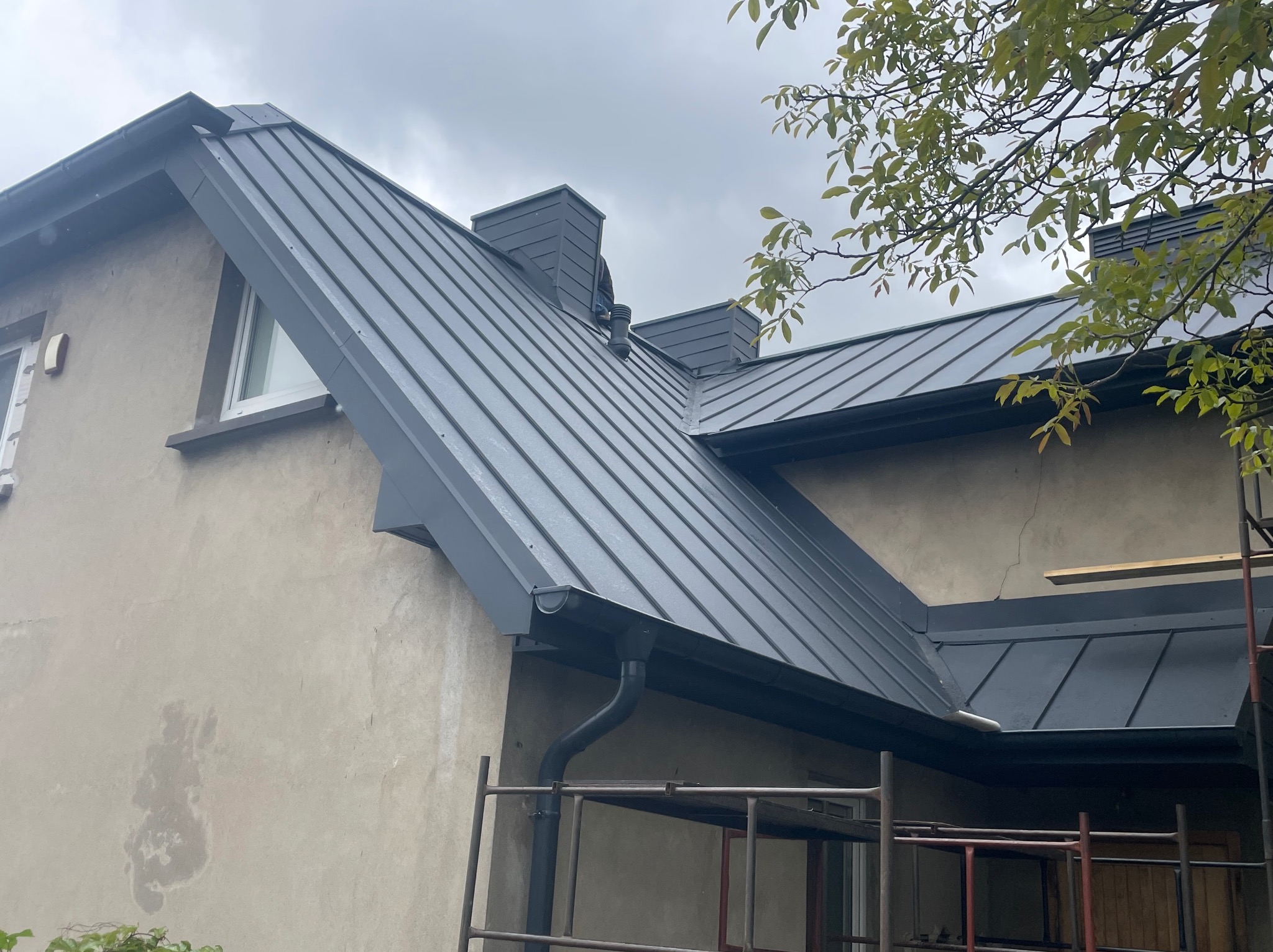 Szary dach z blachy trapezowej na budynku z beżową elewacją. Widoczne rynny, kominy wentylacyjne i rusztowanie. Fragment drzewa z liśćmi w prawym górnym rogu.