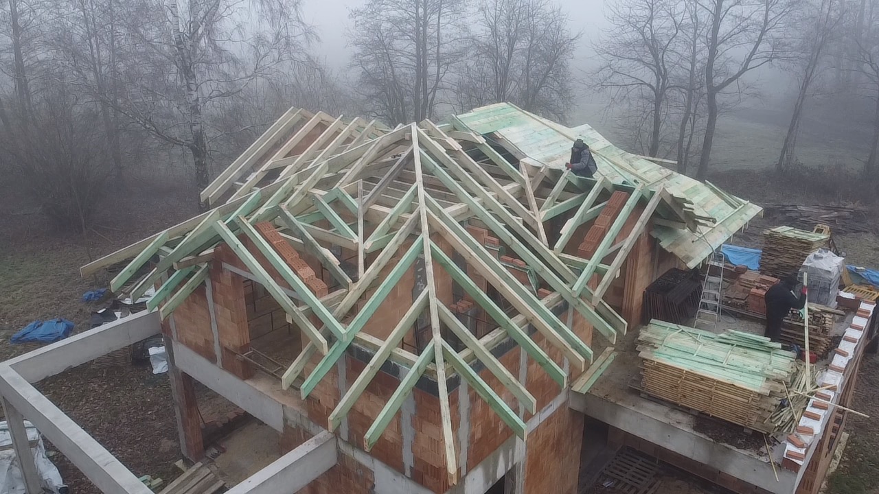 Konstrukcja dachu z drewnianymi belkami na budynku z cegły, widoczne przygotowane deski, praca w toku, mglista pogoda w tle, częściowo pokryty dach.