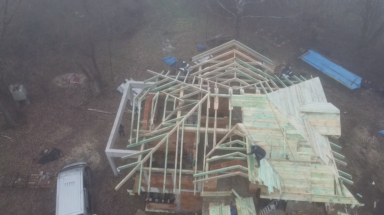 Budowa domu w trakcie realizacji, widok z góry. Widoczne drewniane elementy konstrukcyjne dachu i ściany z cegieł. Na zewnątrz budynku leżą materiały budowlane.