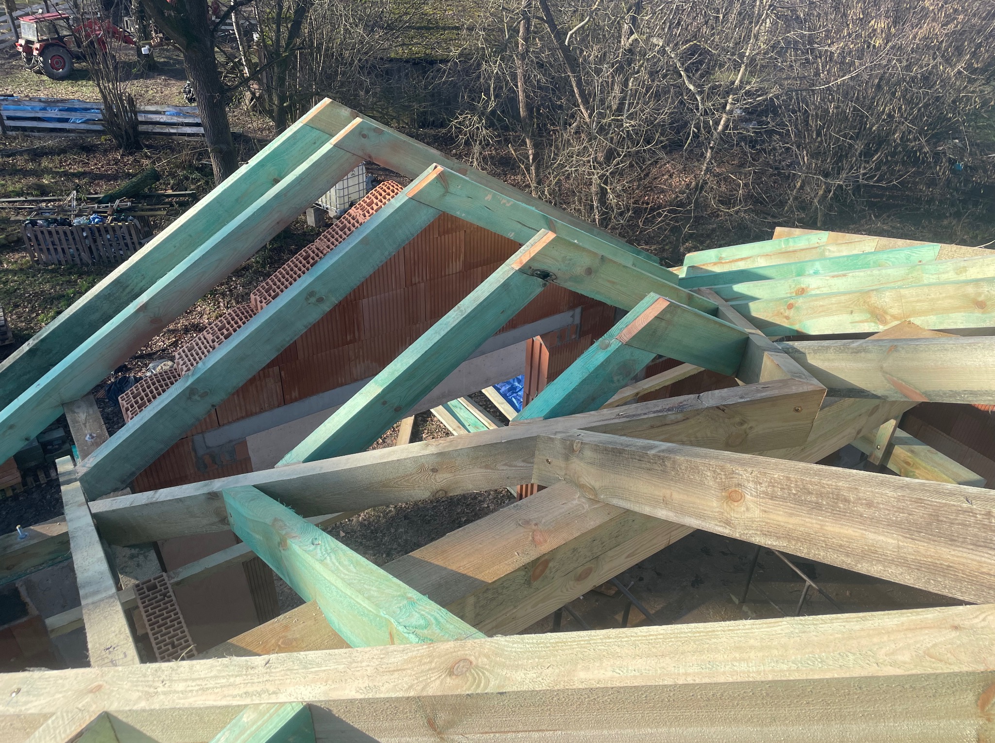 Konstrukcja drewniana dachu w trakcie budowy, zaimpregnowane elementy na tle cegieł i drzew. Widok z góry na elementy więźby dachowej.