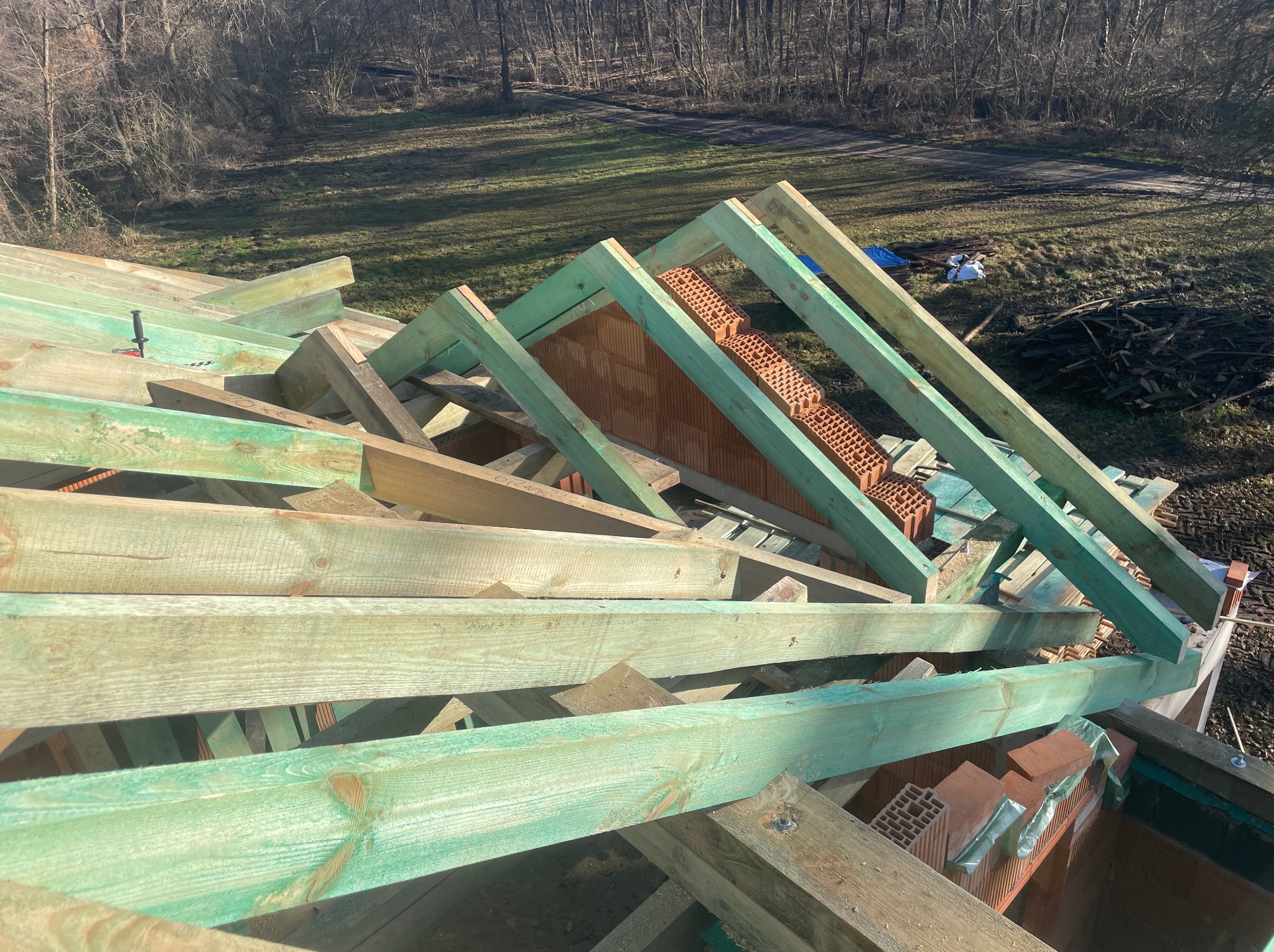 Konstrukcja dachu z impregnowanego drewna i cegły ceramicznej, widok z góry, w tle drzewa i trawa. Etap budowy dachu z użyciem drewna o zielonkawym zabarwieniu.