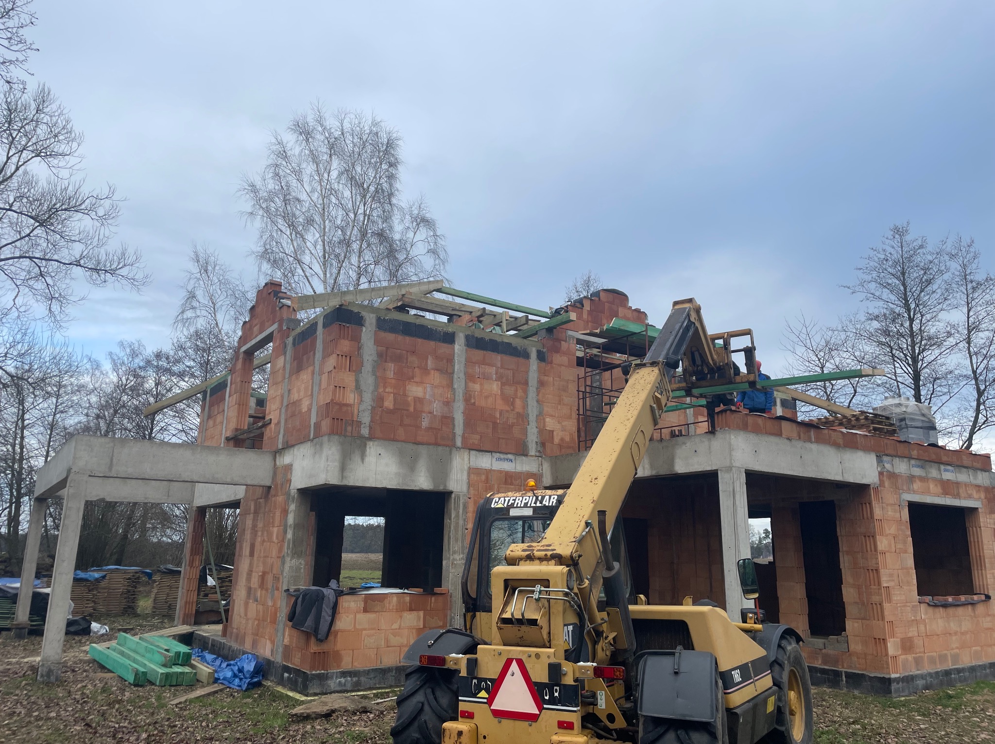 Budowa domu z czerwonej cegły, widoczny podnośnik Caterpillar i elementy konstrukcji dachu w trakcie montażu. Prace dekarskie w toku, widok na surowy stan budynku.