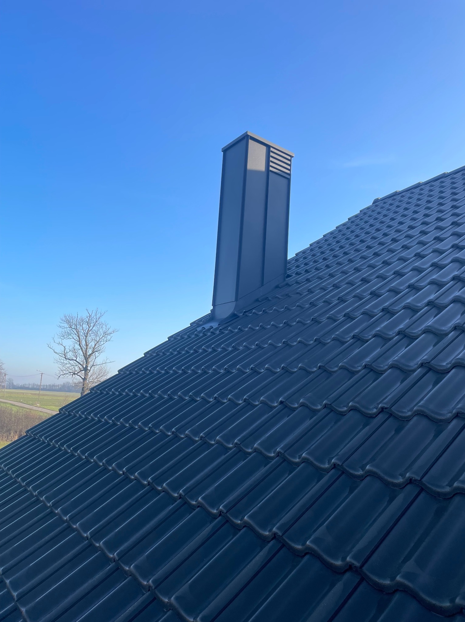 Ciemna dachówka ceramiczna z kominem wentylacyjnym na tle błękitnego nieba i odległego krajobrazu. Widok z góry, ostre światło słoneczne.
