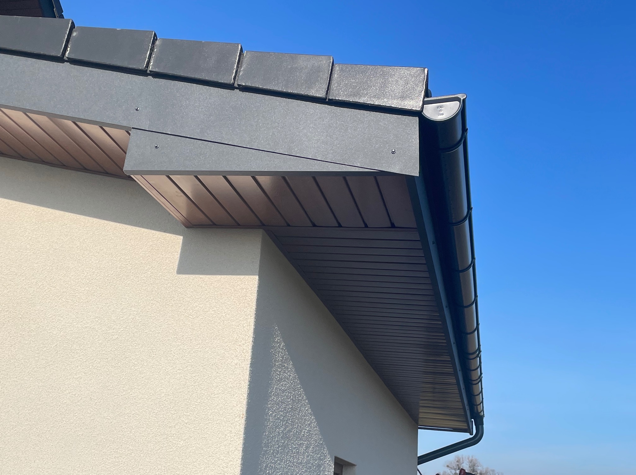 Szczegół dachu z ciemnoszarą dachówką, obróbką blacharską i rynną, widoczne podsufitka i fragment ściany budynku na tle błękitnego nieba.