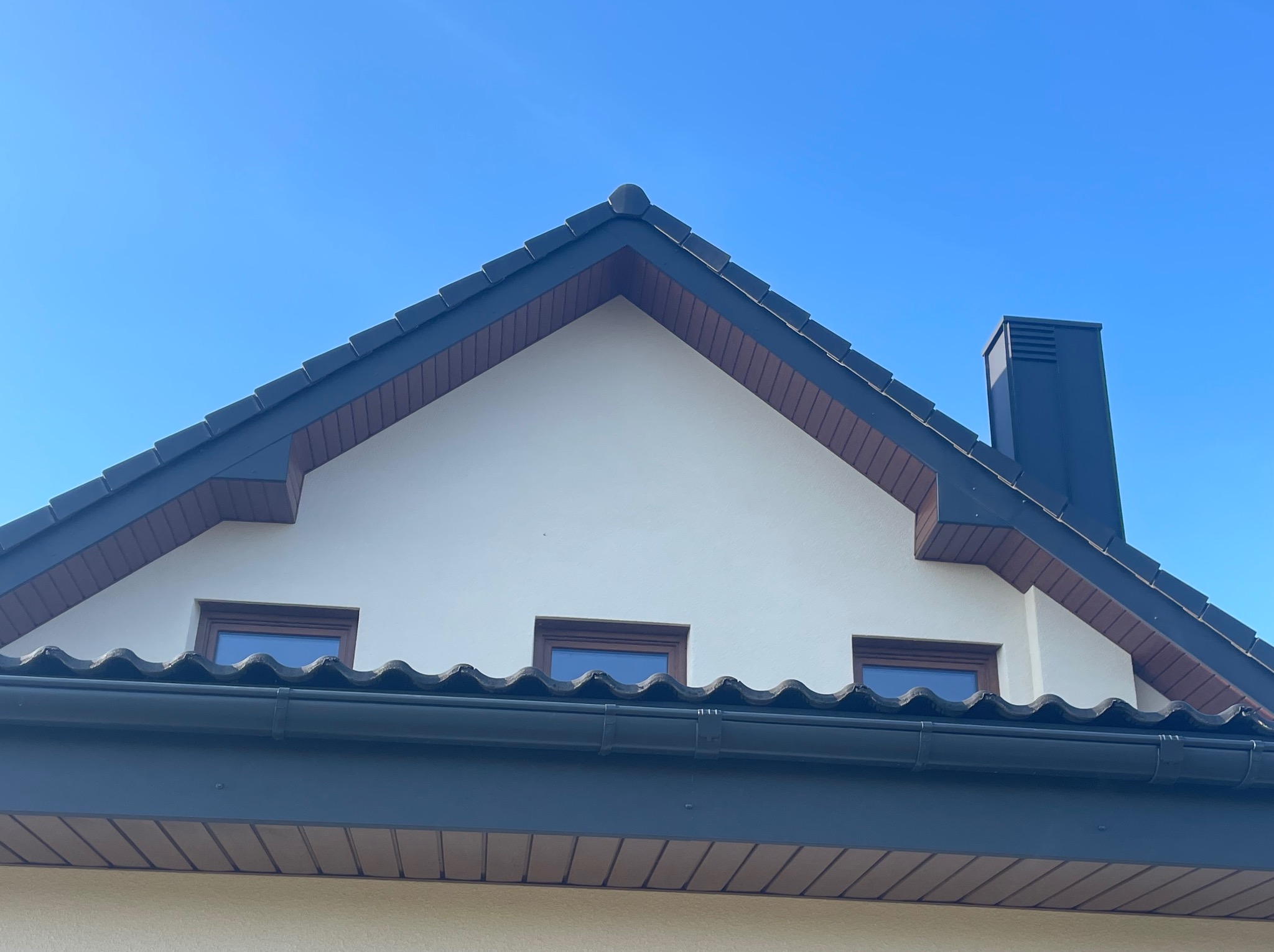 Szczyt domu z ciemną dachówką falistą, trzema oknami i ciemną rynną na tle błękitnego nieba, widok z dołu.