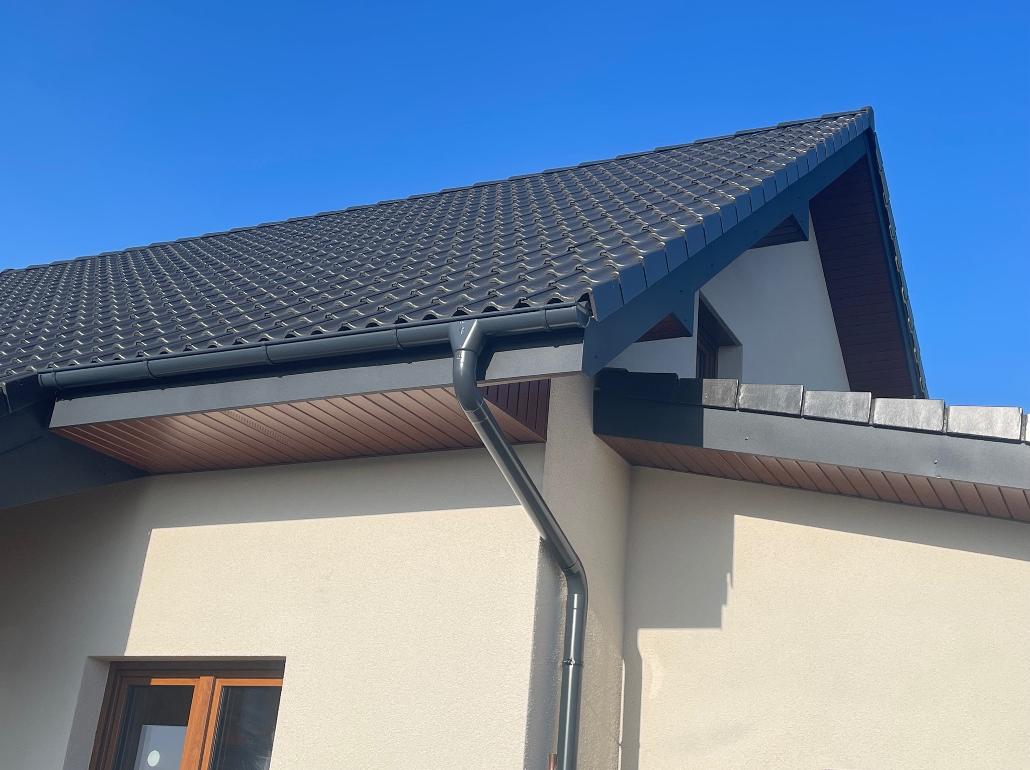Szary dach z dachówki ceramicznej i system rynnowy w kolorze antracytowym na jasnym budynku w słoneczny dzień. Widoczny fragment okna.