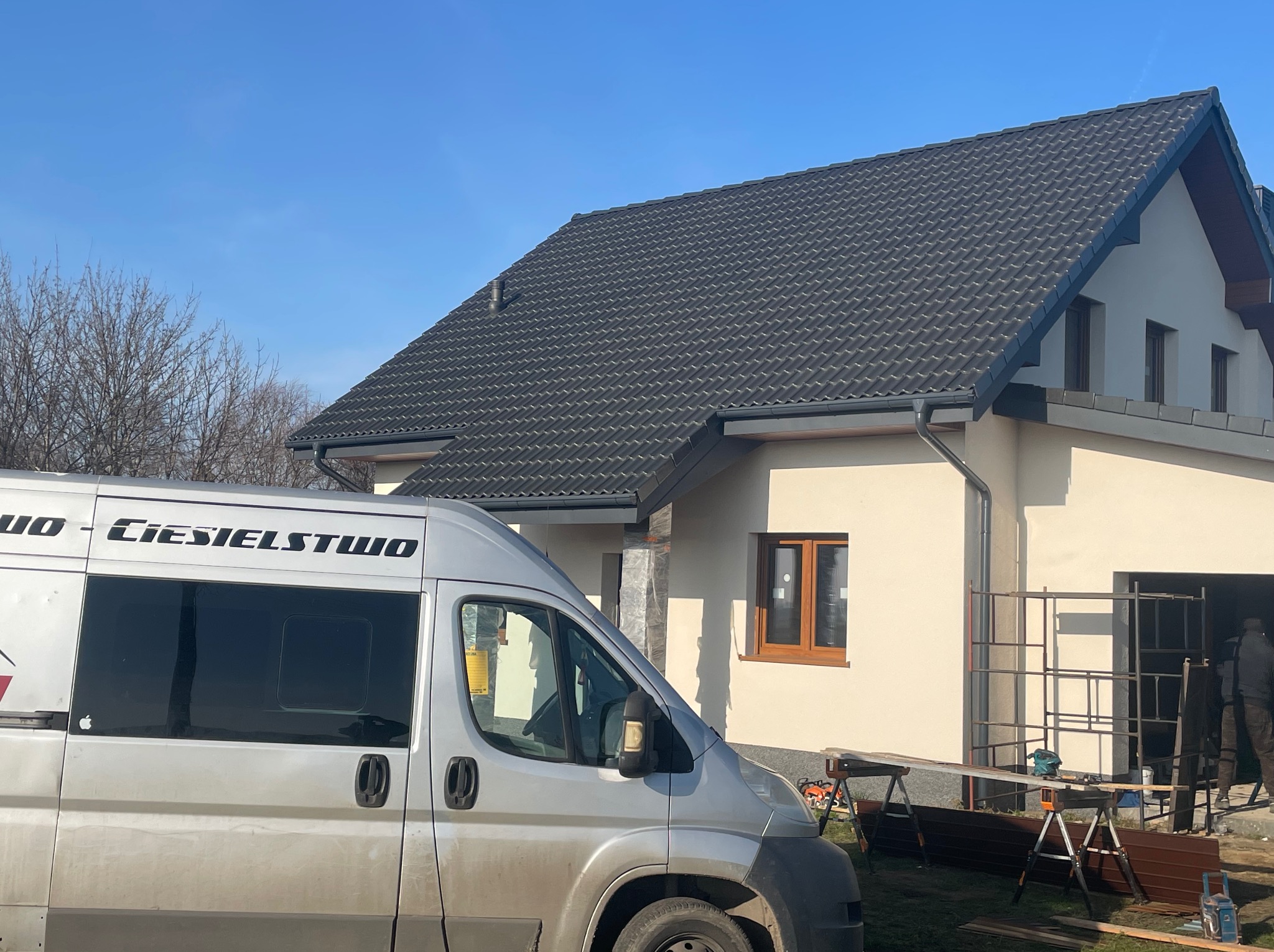 Dom z grafitowym dachem z dachówki ceramicznej i zamontowanymi rynnami. Na pierwszym planie bus z napisem 'Ciesielstwo'.