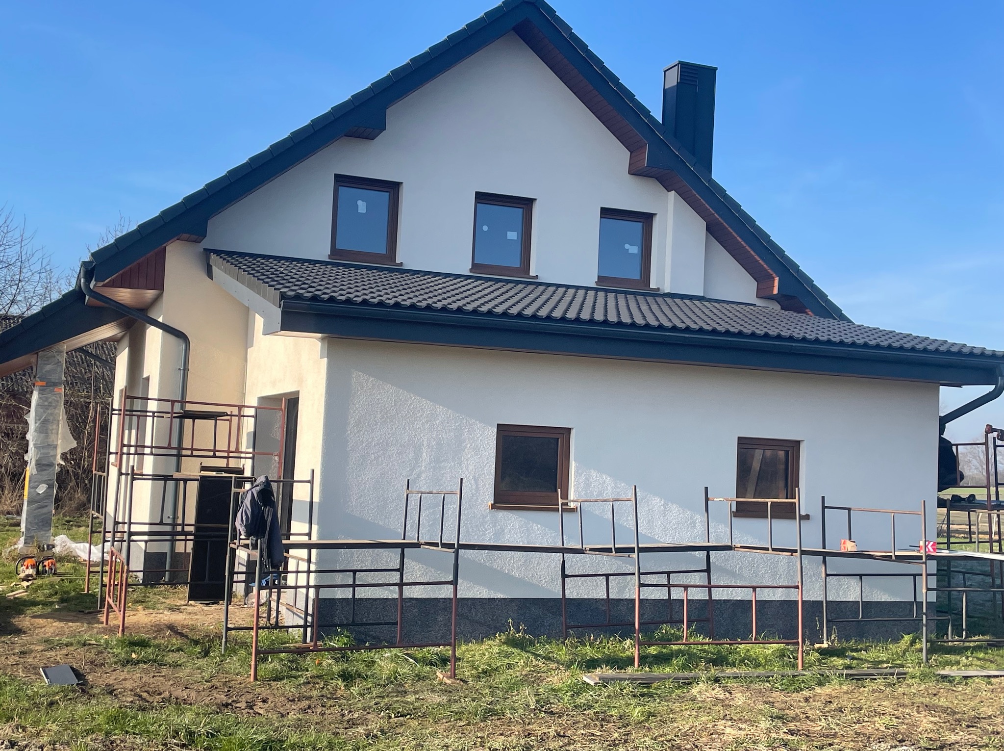 Nowy dom jednorodzinny z ciemną dachówką i rynnami, widoczny fragment rusztowania. Jasna elewacja i okna w brązowych ramach, błękitne niebo w tle.