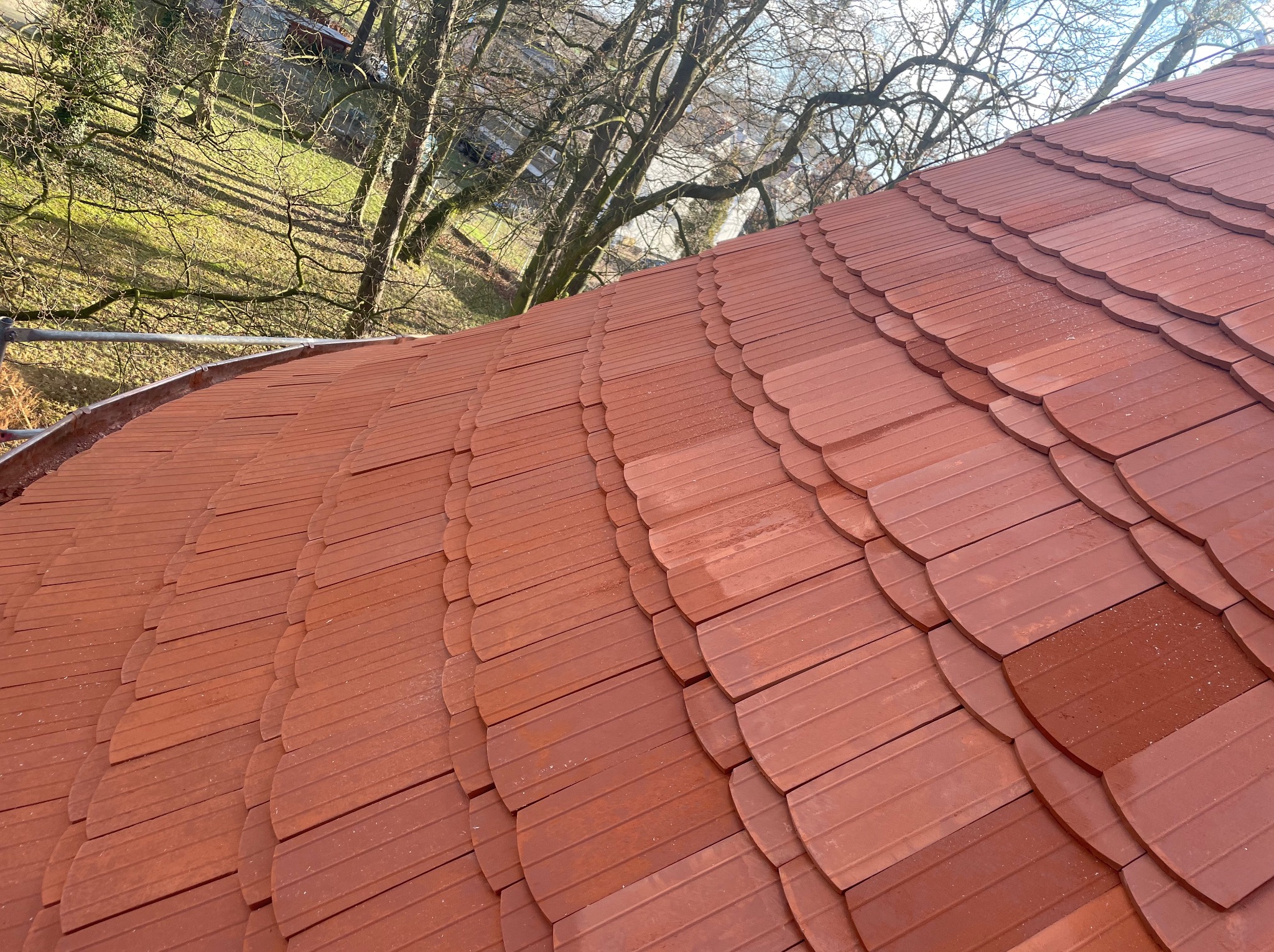Czerwona dachówka karpiówka na zakrzywionym dachu, widok z góry, z elementami orynnowania i drzewami w tle. Układ dachówek tworzy wzór łuski.