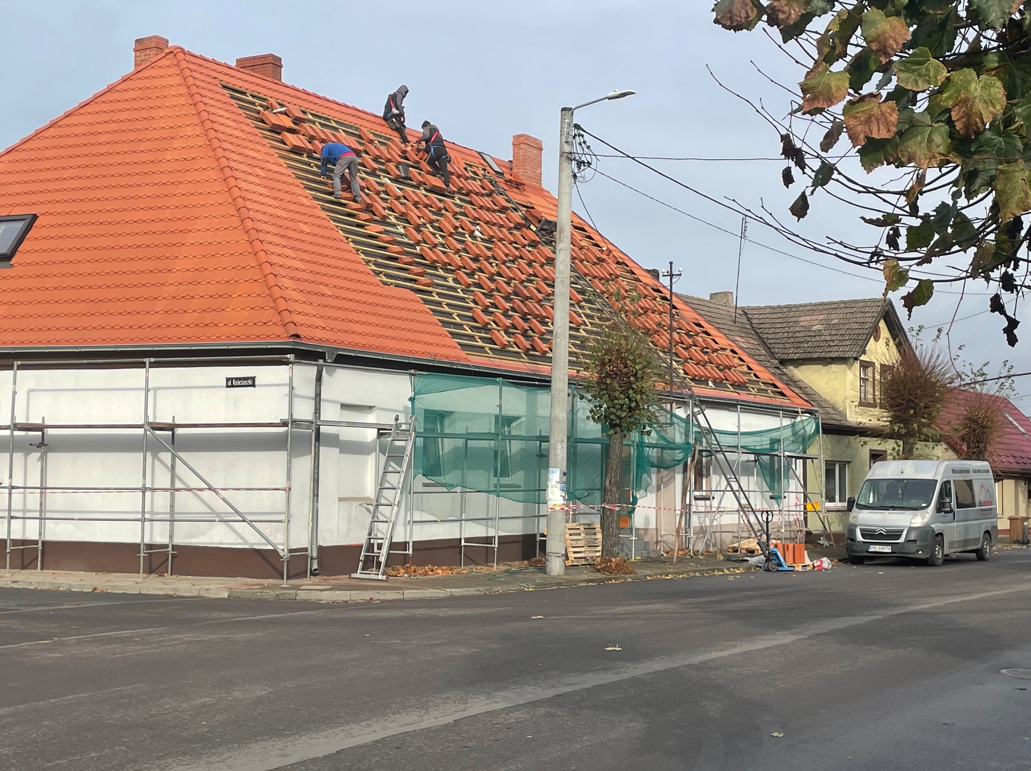 Ekipa dekarzy układa czerwoną dachówkę na konstrukcji dachu domu. Widoczne rusztowania i samochód dostawczy na ulicy. Prace dekarskie w toku.