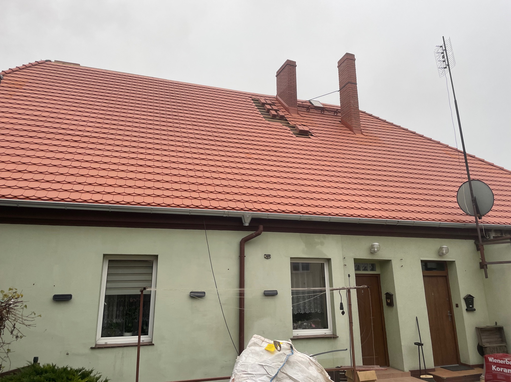 Ceglasty dach z uszkodzeniami w pobliżu komina na budynku w Dobrzycy. Widoczne rynny, antena satelitarna. W tle pochmurne niebo.