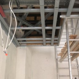 Usługi Budowlane Dariusz Lewandowski - Montaż metalowego rusztu sufitu podwieszanego z widocznymi przewodami elektrycznymi, drabina oparta o ścianę, surowy stan wykończenia wnętrza.
