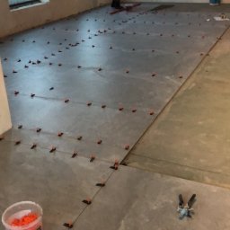 Usługi Budowlane Dariusz Lewandowski - Układanie płytek wielkoformatowych na podłodze z użyciem systemu poziomowania płytek, widoczny pracownik w tle i wiadro z klinami.