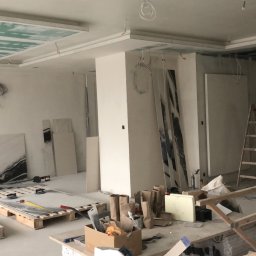 Usługi Budowlane Dariusz Lewandowski - Wnętrze w trakcie remontu: sufit z płyt gipsowo-kartonowych z widoczną siatką spoin, duże płyty imitujące marmur oparte o ścianę i leżące na paletach, drabiny i rusztowanie, widoczne przewody...