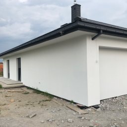 Usługi Budowlane Dariusz Lewandowski - Biała elewacja domu z ciemnymi oknami i dachem, widoczna osoba pracująca przy wykończeniu, teren wokół domu w trakcie prac budowlanych.