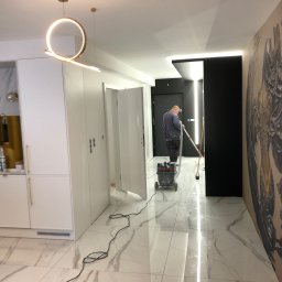 Usługi Budowlane Dariusz Lewandowski - Wnętrze mieszkania w trakcie prac wykończeniowych: mężczyzna szlifuje podłogę polerką, widoczny sprzęt do polerowania, marmurowa podłoga, designerska lampa i grafika na ścianie.