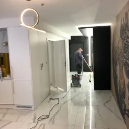Wnętrze mieszkania w trakcie prac wykończeniowych: mężczyzna szlifuje podłogę polerką, widoczny sprzęt do polerowania, marmurowa podłoga, designerska lampa i grafika na ścianie.