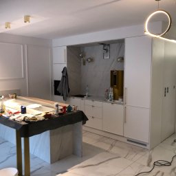 Usługi Budowlane Dariusz Lewandowski - Wnętrze kuchni w trakcie remontu: marmurowa podłoga i wyspa przykryta folią, białe szafki z złotymi uchwytami, wisząca lampa w kształcie okręgu, ściany przygotowywane do malowania.