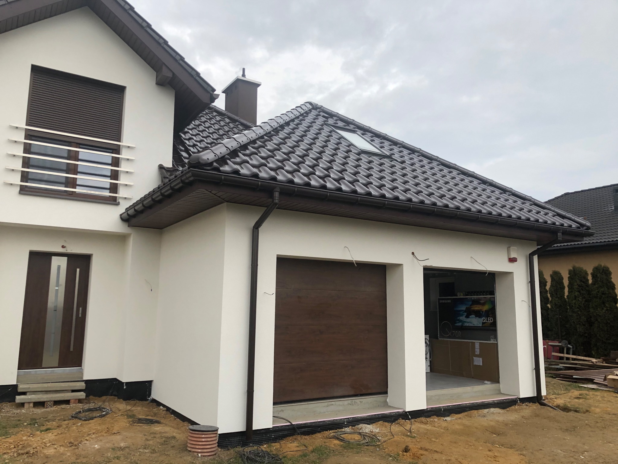 Wykończona elewacja domu jednorodzinnego w Żorach z brązową stolarką okienną i garażową oraz dachem z ciemnej dachówki ceramicznej, widoczne niedokończone otoczenie budynku.